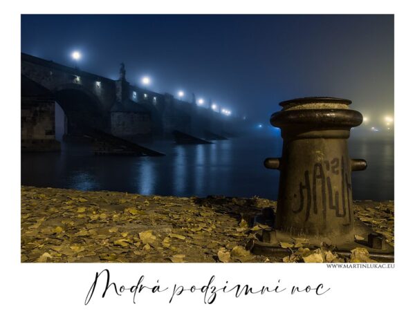 Modrá podzimní noc - Noční fotografie Karlova mostu s osvětlenými oblouky, modře tónovaná scéna, v popředí bollard a padající listí, autor Martin Lukač.