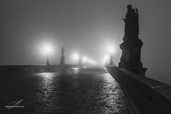 Noční Karlův most zalitý mlhou a světlem luceren, s dramatickými siluetami soch, autor Martin Lukač, černobílá
