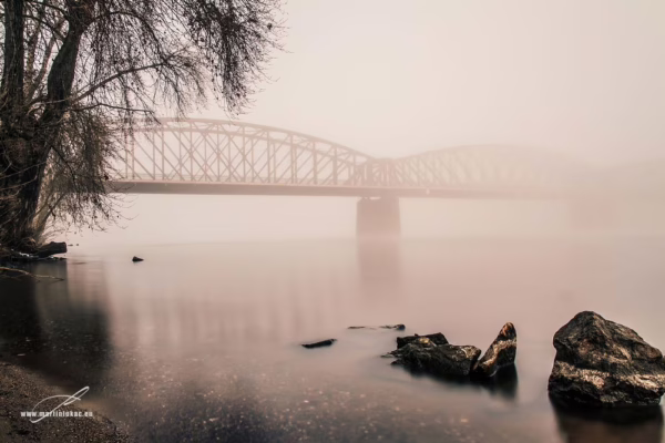 Černobílá fotografie železničního mostu přes Vltavu v ranní mlze, chladná atmosféra a klidná voda, autor Martin Lukač
