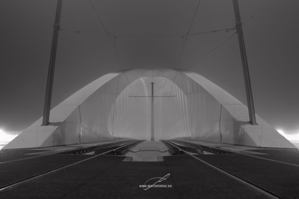 Černobílá fotografie Trojského mostu v mlze s tramvajovými kolejemi a osvětlenou konstrukcí, autor Martin Lukač, černobílý