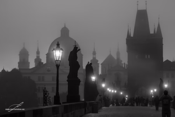 Karlův most za večerní modré hodiny s pouličními lampami a postavami, autor Martin Lukač, černobílá